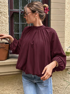 A woman wearing a loose-fitting, long-sleeve burgundy blouse with a gathered high neck and elasticized cuffs, paired with light blue jeans, standing outdoors next to a brown leather handbag.