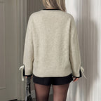 A woman shown from the back wearing an oversized cream-colored knit sweater with black trim at the collar and cuffs, plus matching black shorts. The sweater has ribbon details at the cuffs.