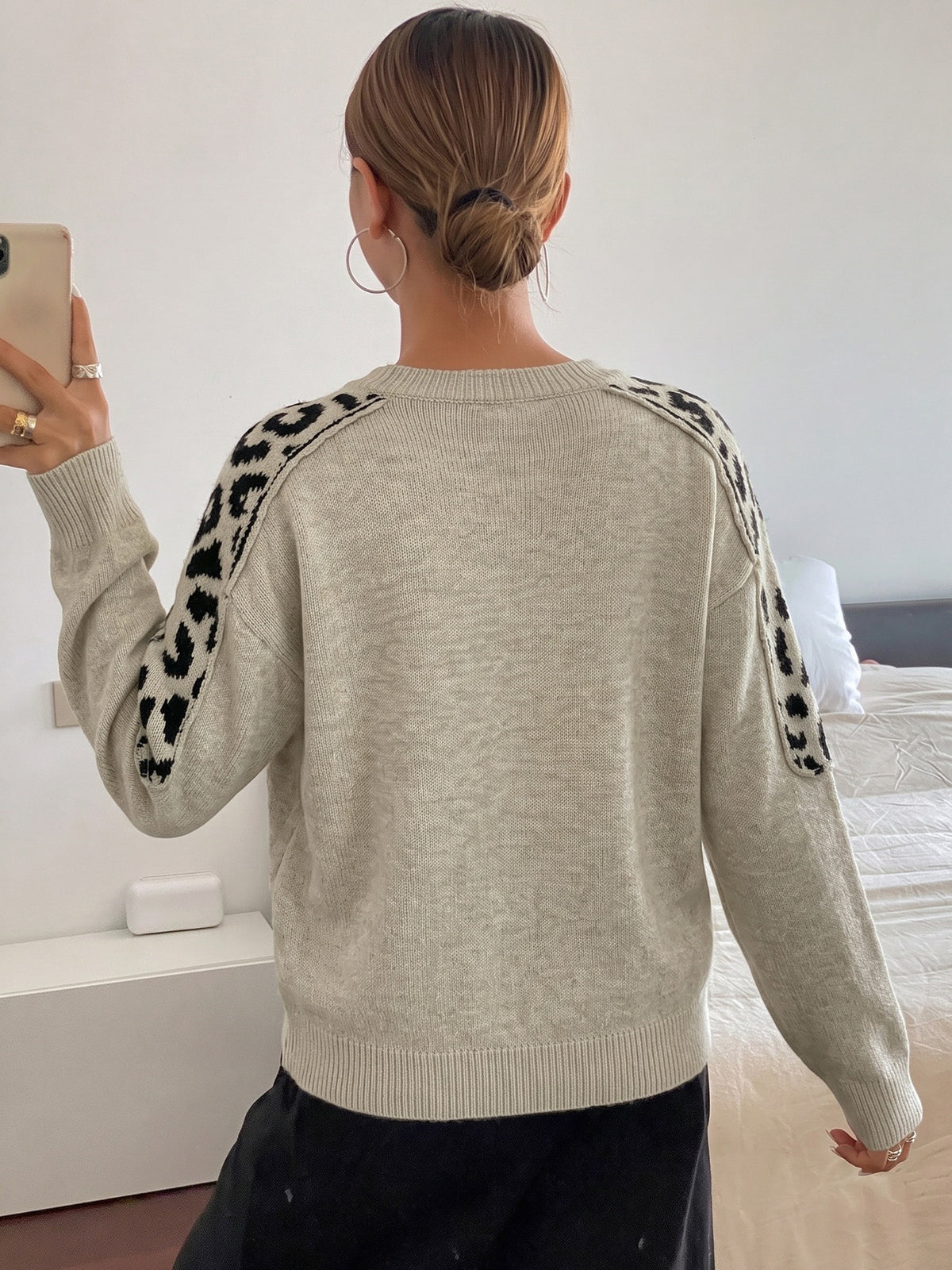 A woman viewed from behind wearing a light beige knit sweater with black leopard-print detailing on the shoulders and upper sleeves, paired with black pants in a bedroom setting.