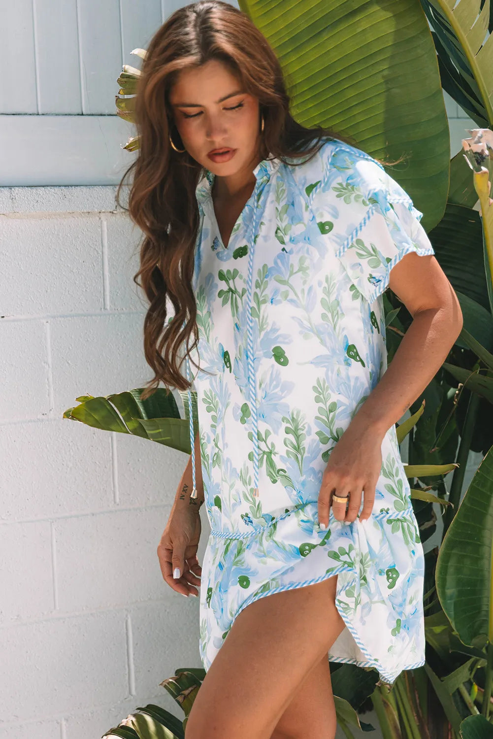 Woman wearing a floral cover-up dress standing in front of a white wall with green plants.